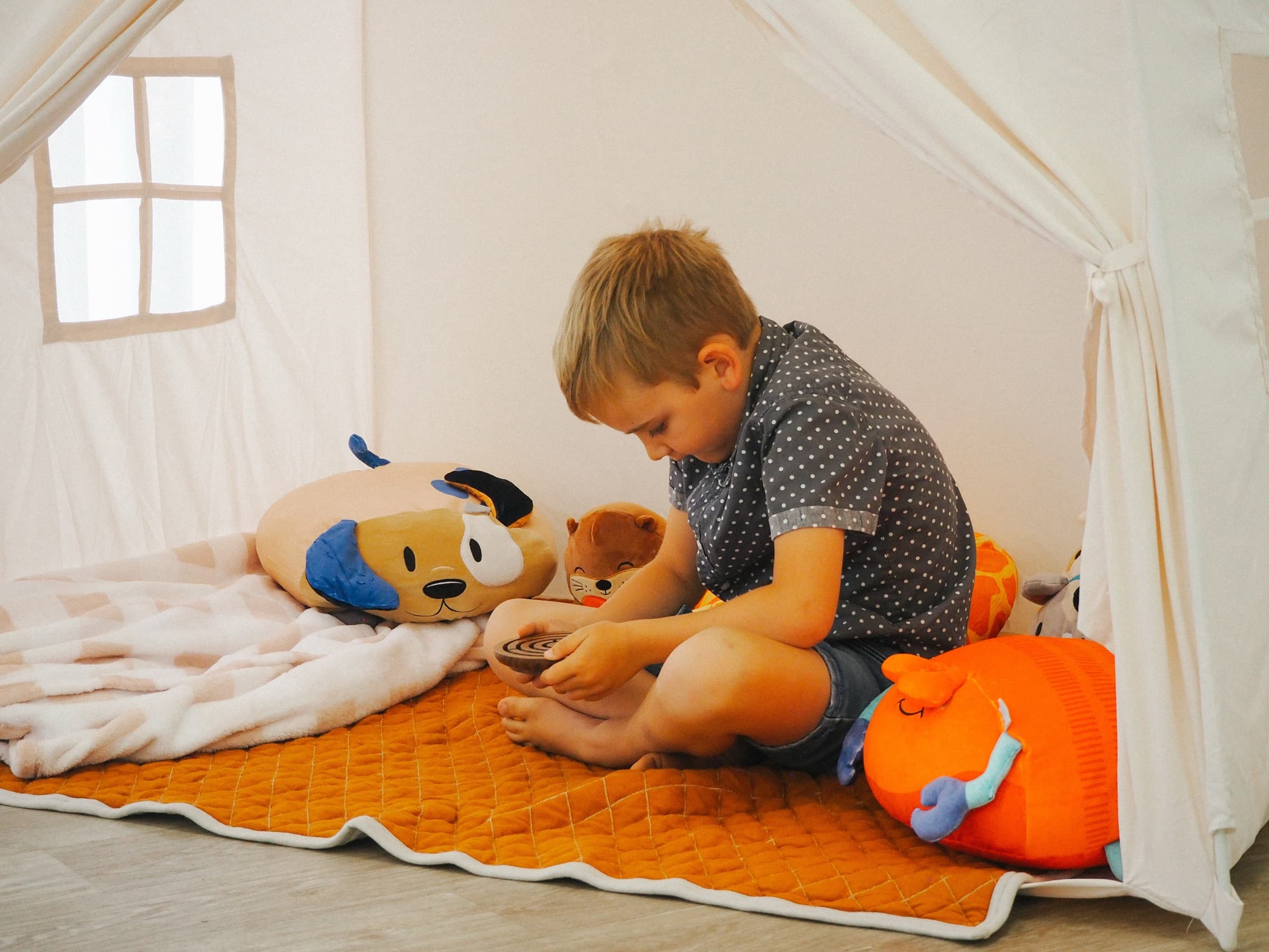 Young boy relaxing inside a cosy play tent surrounded by stuffed animals and soft cushions