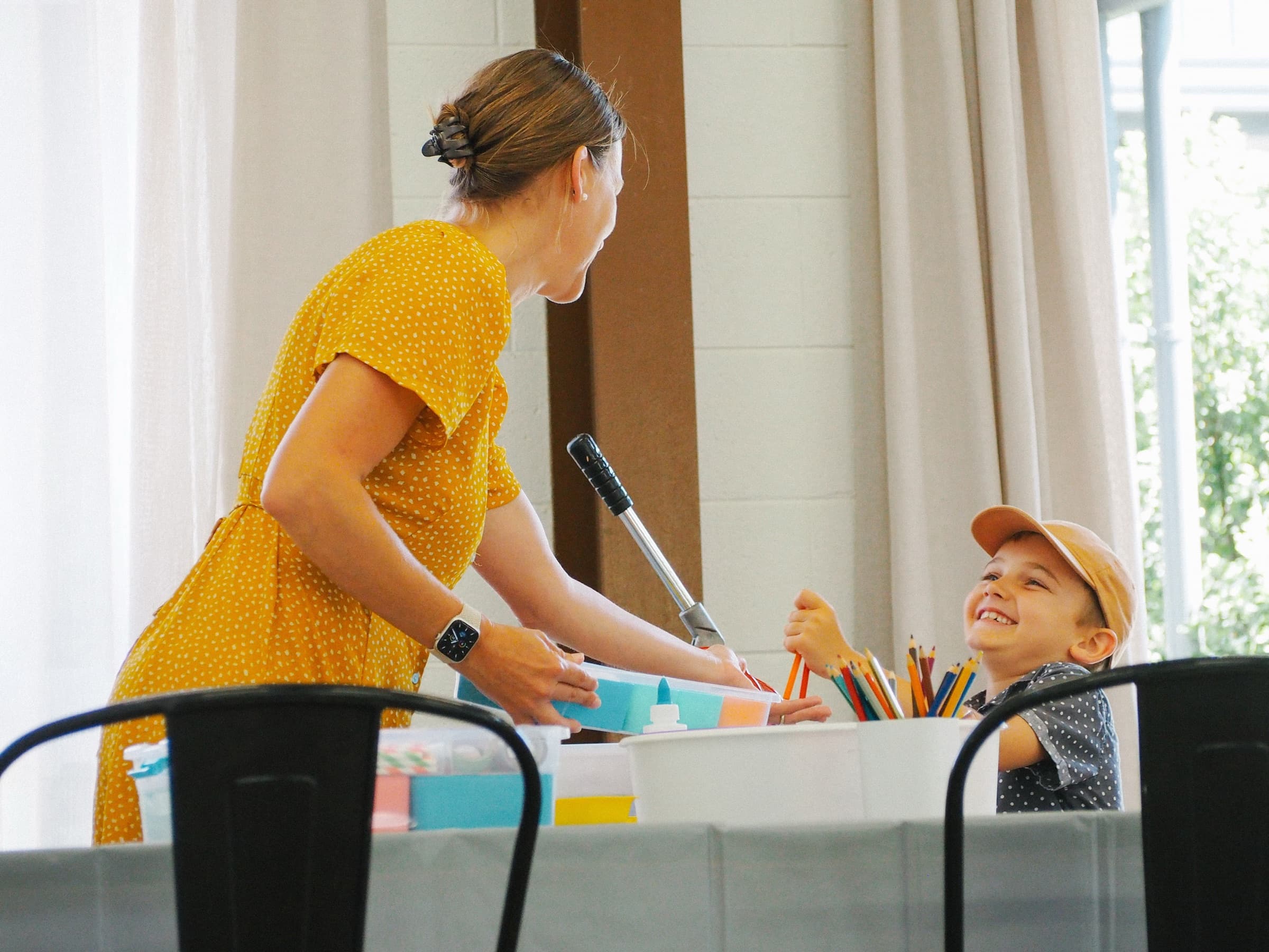 Event Sitter in a yellow dress helping a smiling young boy at a craft table
