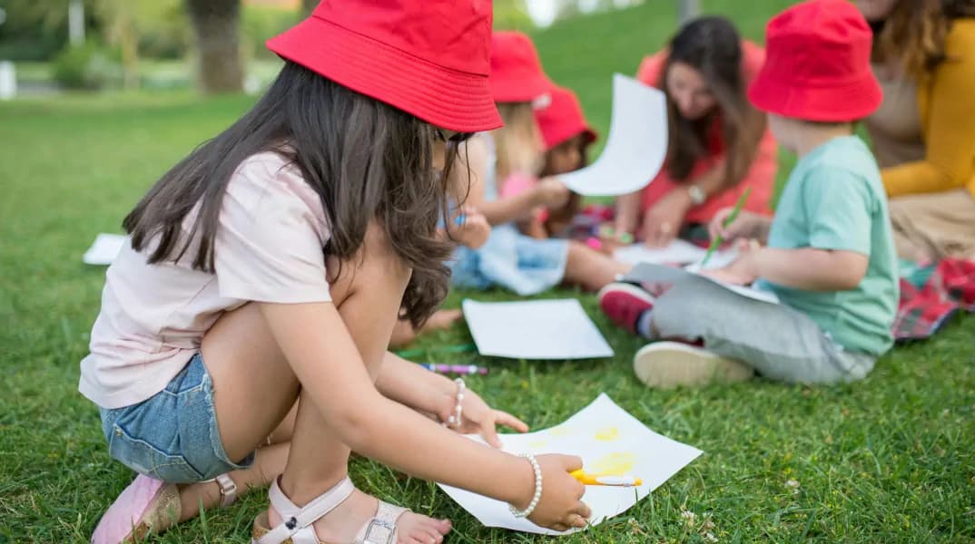 Children sitting on grass in an outdoor setting, engaged in drawing and crafting activities.