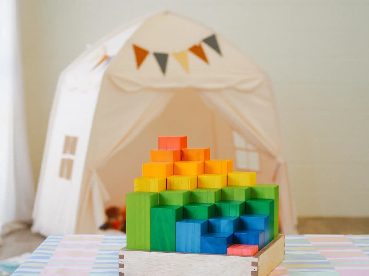 Rainbow-coloured wooden building blocks stacked in a pyramid in front of a cosy play tent with bunting flags