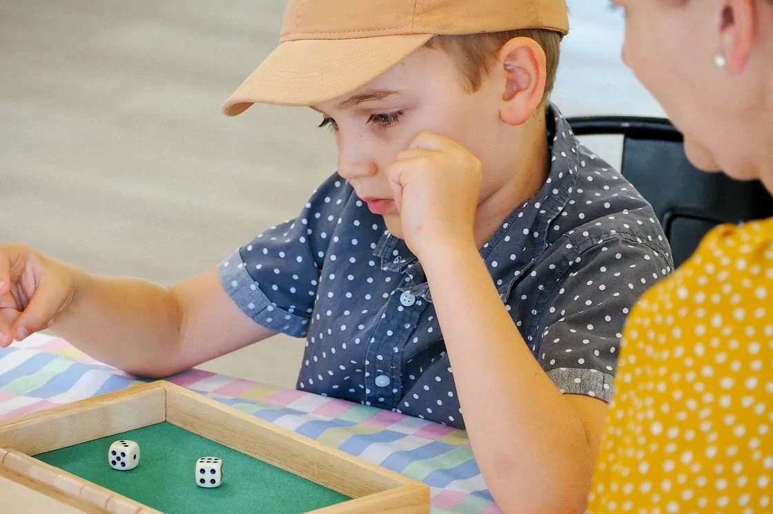 A young boy in a polka-dotted shirt and beige cap is deep in concentration while playing a dice game in a wooden tray.