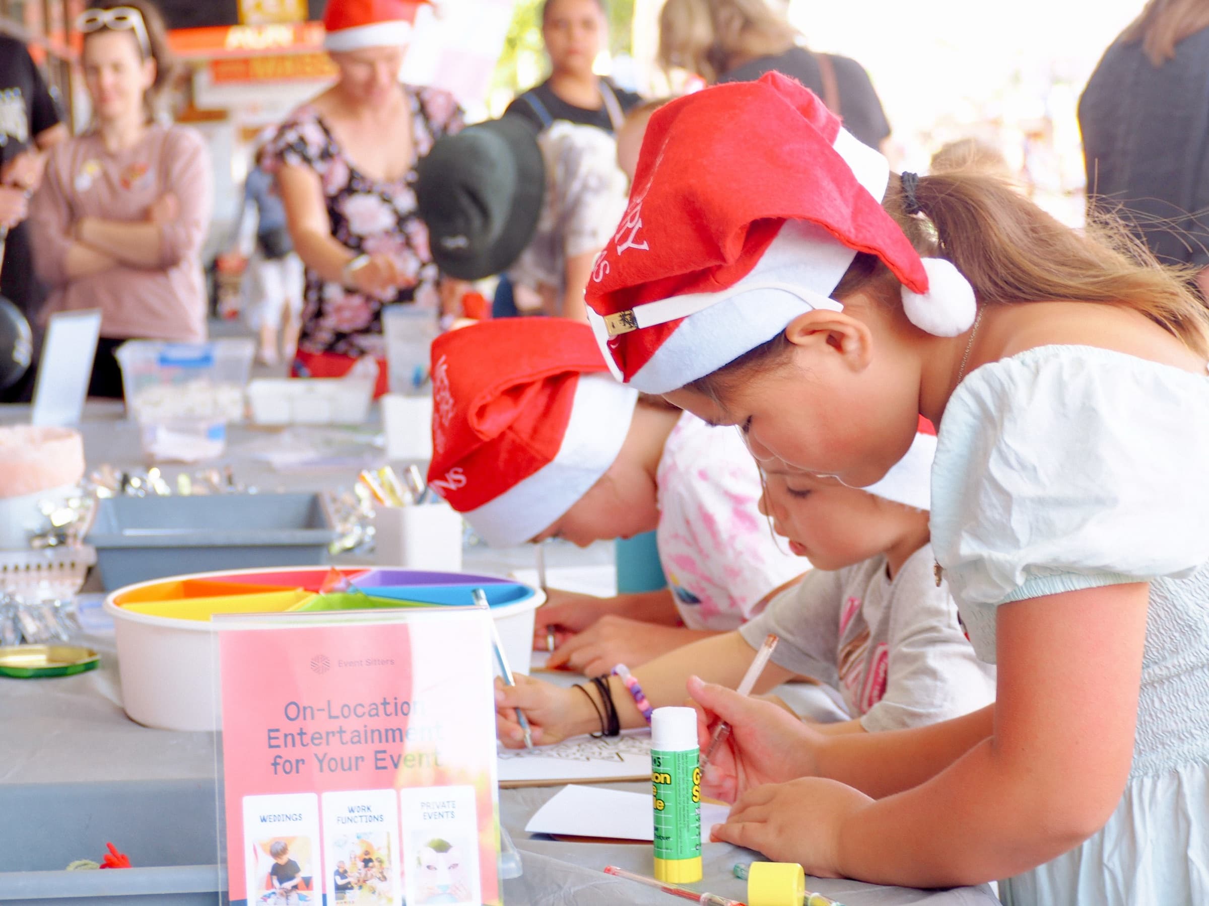 Children in Santa hats doing arts and crafts at an outdoor public event, with Event Sitters signage visible