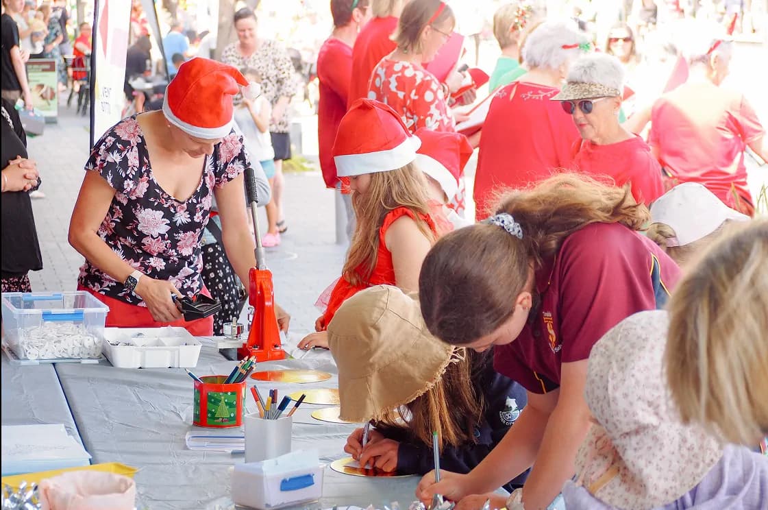 A group of children and adults gather around a table at an outdoor event, engaged in holiday crafts.